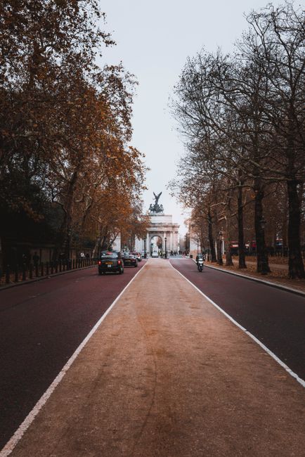 A wide city street with a central beige-colored pedestrian pathway made of textured material, flanked by dark asphalt lanes on both sides, leading towards a large arch monument topped with an equestrian statue. On the left side, there are parked cars, and on the right, a person riding a bicycle. Tall trees with mostly bare branches line both sides of the street, some with brown leaves still clinging to the branches, indicating late autumn or early winter. The scene is illuminated by diffused natural daylight under an overcast sky, creating a neutral, subdued atmosphere. In the distance, the arch and statue serve as a focal point, with the surrounding environment suggesting a prominent urban park setting. This scene, associated with private or alternative waste disposal, emphasizes the importance of organized rubbish collection in public spaces, as observed by the absence of visible waste or debris along the pathway and street edges, aligning with professional rubbish removal services like those offered by Rubbish Collection Bayswater.