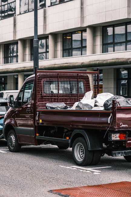 A small, dark red flatbed truck parked on an urban street in front of a modern building with large, reflective glass windows and a light-colored, concrete exterior. The truck's bed is filled with various types of rubbish, including black and white plastic bags as well as larger bulky items, suggesting an on-site clearance or private waste removal. A metal mesh panel forms a partial barrier at the truck's cab, providing some separation between the driver's area and the waste load. The scene is set during daylight hours, with natural light illuminating the vehicle and surroundings. Nearby, there are additional parked vehicles and a street lamp, emphasizing the urban environment. The image is relevant to independent waste collection and rubbish removal services, such as those offered by Rubbish Collection Bayswater, supporting the concept of alternative waste handling solutions outside of local authority disposal channels.