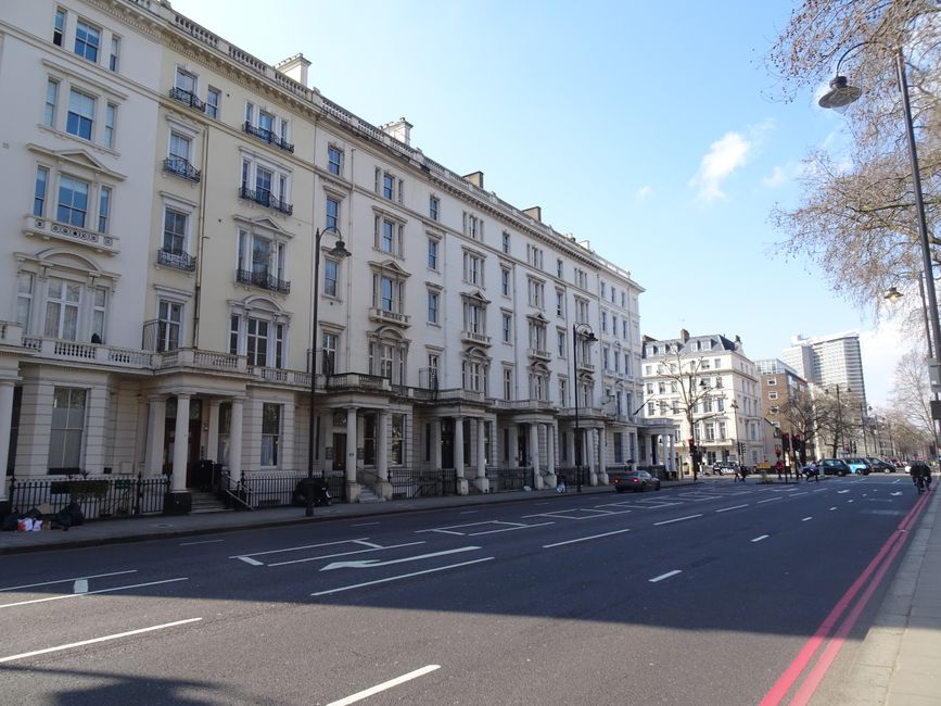 A wide street scene featuring a row of elegant, white, multi-story Victorian-style terraced buildings with ornate architectural details, such as decorative cornices and small balconies with black wrought-iron railings. The buildings have large sash windows, some with bay windows, and stone steps leading to dark-colored front doors. The street is paved with asphalt and has marked parking bays along the curb, with a few parked cars visible. On the right side of the image, a leafless tree extends its branches toward the clear blue sky, which has a few scattered white clouds. A street lamp with a curved arm is also visible near the tree. The overall scene is well-lit, suggesting a sunny day, and the street appears quiet with minimal pedestrian activity, with some small items or bags placed near the base of the buildings, indicating recent waste collection or disposal activities. The environment suggests a typical urban residential area with proximity to private waste handling services, such as those offered by Rubbish Collection Bayswater, for alternative rubbish removal in the area.