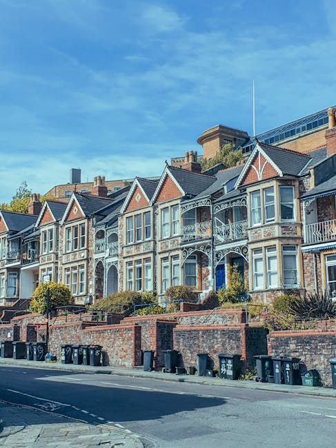 A row of traditional Victorian-style terraced houses with ornate white ironwork balconies and decorative trims, constructed from red brick with contrasting white window frames and gables, situated on a residential street. The front gardens are enclosed by low brick walls, with bushes and small trees lining the pavement. In the foreground, a series of black wheelie bins are positioned along the sidewalk, indicating waste collection activity. The scene is captured during daylight hours under a clear blue sky, with some modern buildings visible in the background behind the historic houses. The overall setting suggests a typical urban neighbourhood where private waste collection services, such as those offered by Rubbish Collection Bayswater, may be utilized as an alternative to council-led rubbish disposal, highlighting a context of on-site clearance and independent rubbish management.
