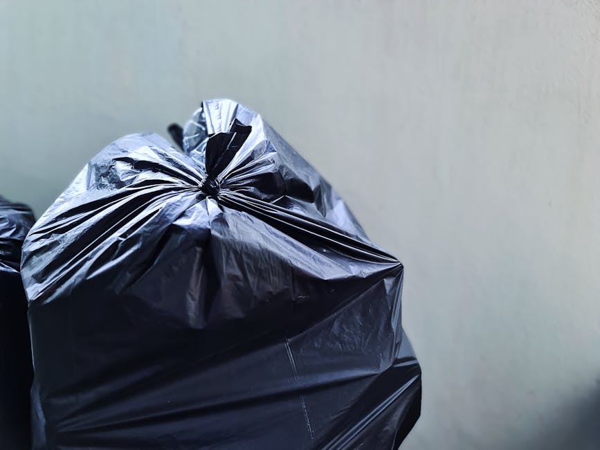 Close-up image of several tightly packed black rubbish bags made of shiny plastic material, with visible creases and folds, placed against a plain, light-colored wall. The bags appear to be filled with waste and are situated on a flat surface, possibly an indoor or outdoor area used for rubbish collection or disposal. The lighting is even and neutral, emphasizing the reflective nature of the plastic bags. The scene suggests a setting related to private waste handling or rubbish removal services, aligned with the context of alternative collection options such as those offered by Rubbish Collection Bayswater. The focus remains on the bags' texture, material, and arrangement, with no other objects or environmental features visible in the frame, supporting an accurate visual description suitable for accessibility and service relevance.